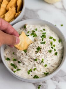 Hand dipping a cracker into a bowl of creamy french onion dip topped with chopped chives and pepper.