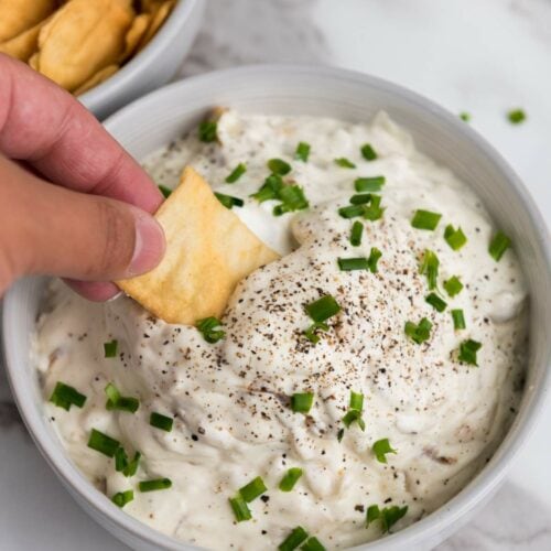 Hand dipping a cracker into a bowl of creamy french onion dip topped with chopped chives and pepper.