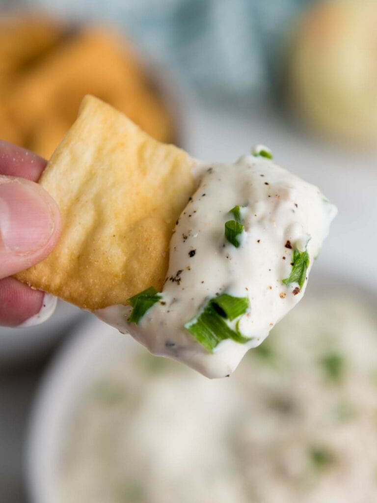 A hand holds a cracker dipped in a creamy french onion sauce with chopped green herbs and pepper.