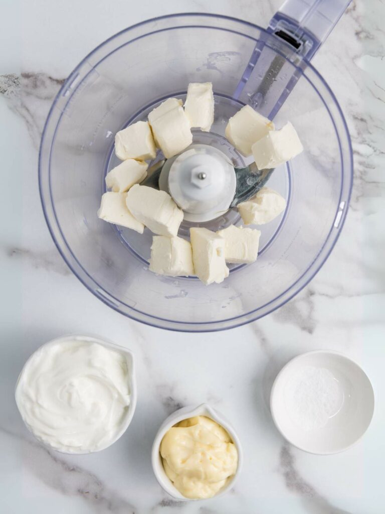 Overhead view of a food processor with cubed cream cheese, next to bowls of sour cream, mayonnaise.