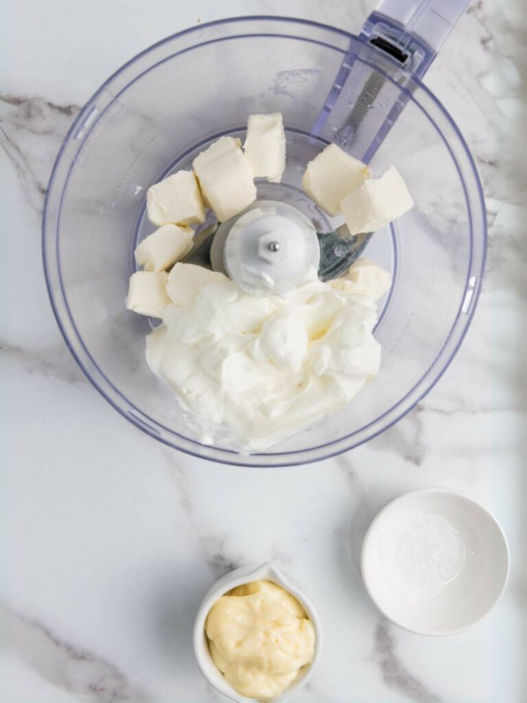 A food processor with cream cheese, sour cream, and mayonnaise. Salt in a small bowl nearby.