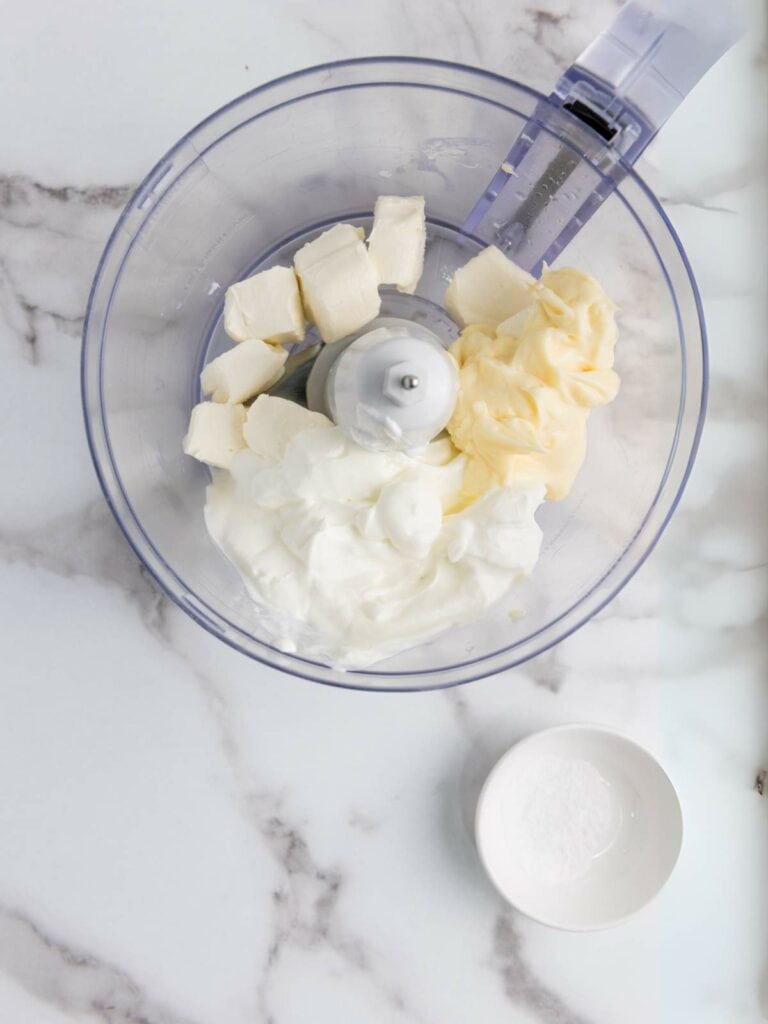 Food processor bowl with cream cheese and sour cream, next to a small dish of salt, on a marble surface.