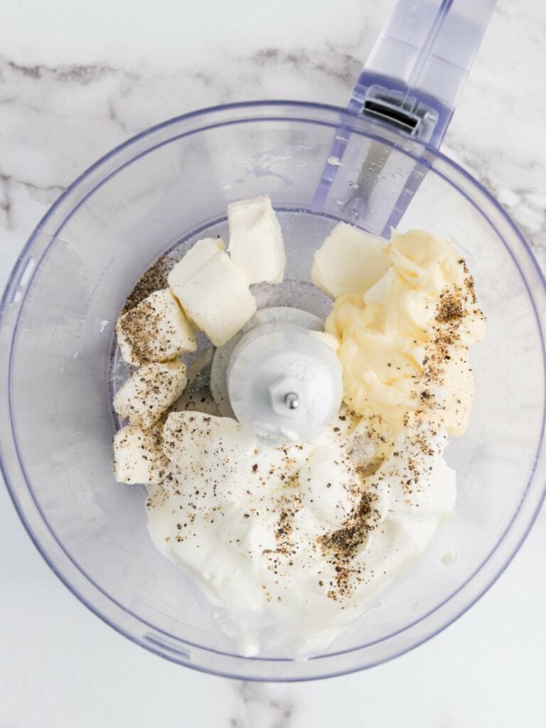 Food processor bowl with butter, mayonnaise, sour cream, and spices on a marble countertop.