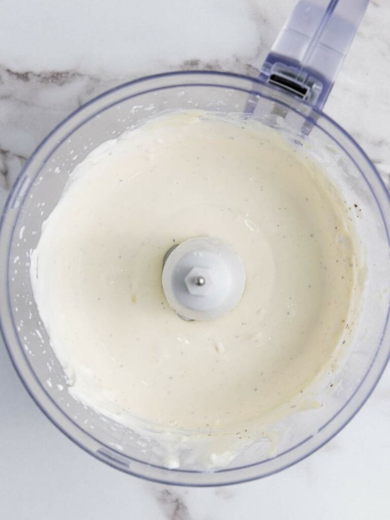 A creamy white sauce is being mixed in a food processor on a marble countertop.