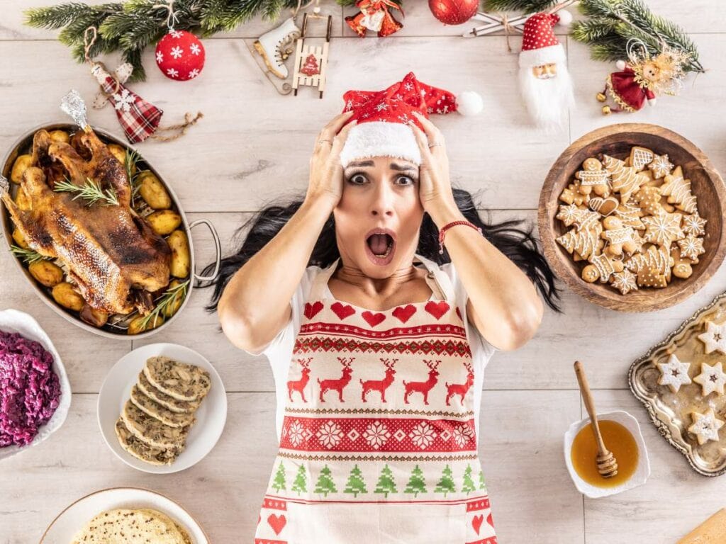 A woman in a festive apron and Santa hat looks surprised while lying on a table surrounded by holiday food and decorations.