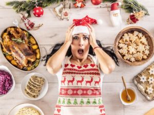 A woman in a festive apron and Santa hat looks surprised while lying on a table surrounded by holiday food and decorations.