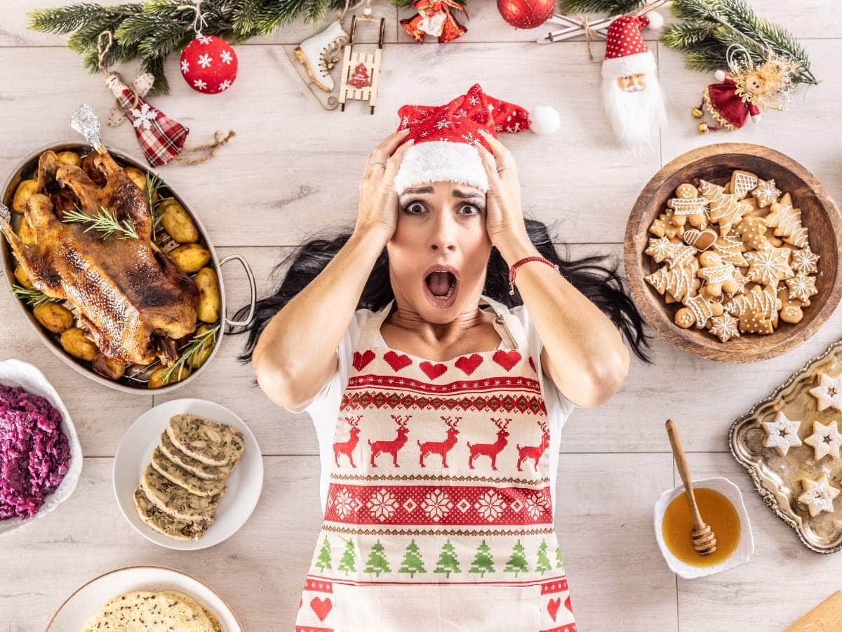 A woman in a festive apron and Santa hat looks surprised while lying on a table surrounded by holiday food and decorations.