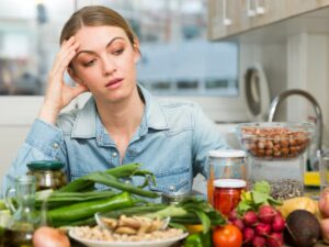 Person in a kitchen surrounded by various fresh vegetables and jars, looking contemplative.