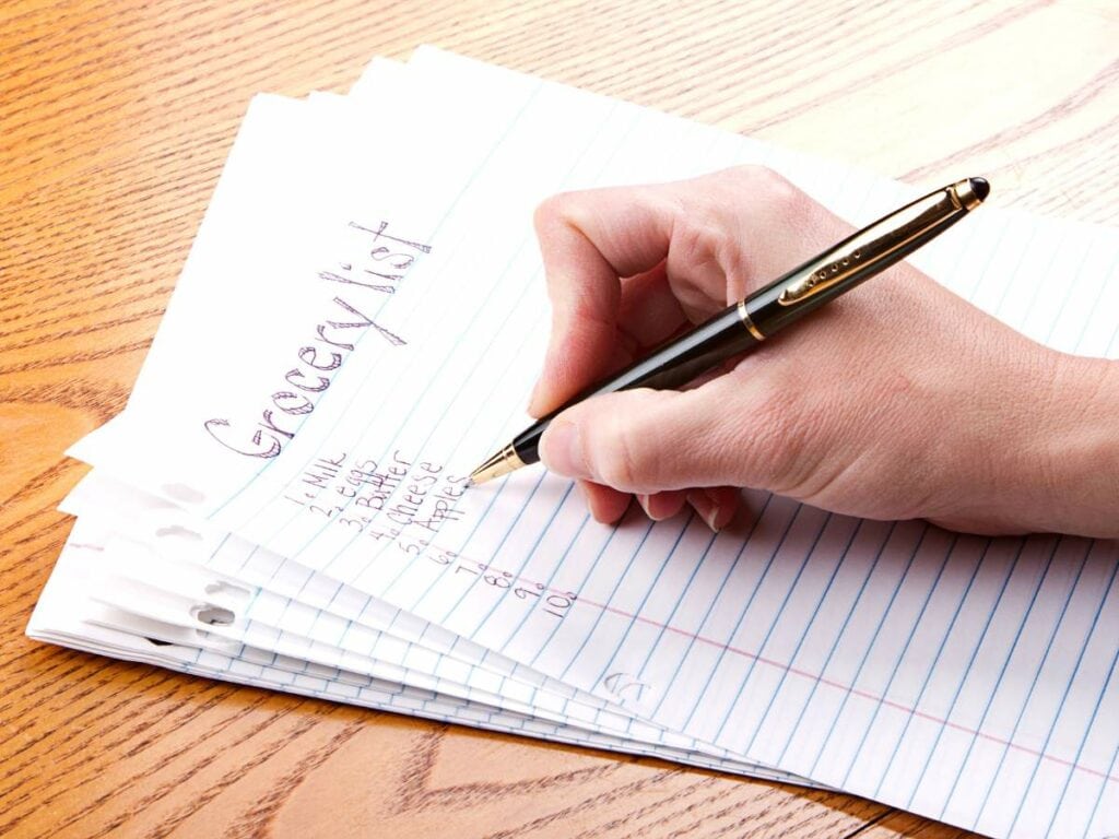 A hand writes a grocery list on lined paper with a pen. Items include milk, eggs, bread, apples, and grapes.