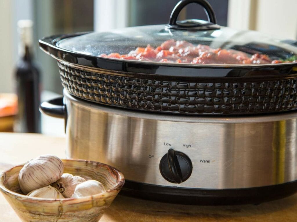 A slow cooker with a clear lid sits on a countertop with a bowl of garlic cloves nearby.