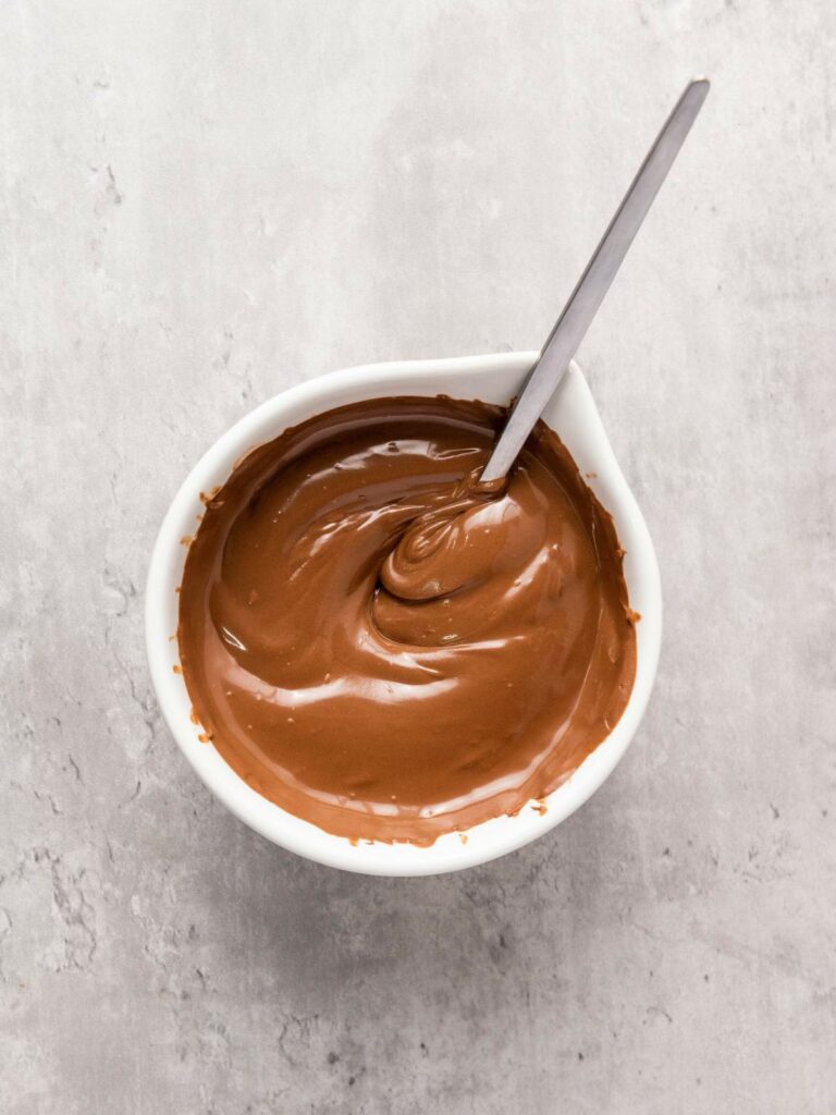 A bowl of creamy chocolate sauce with a spoon on a light gray surface.