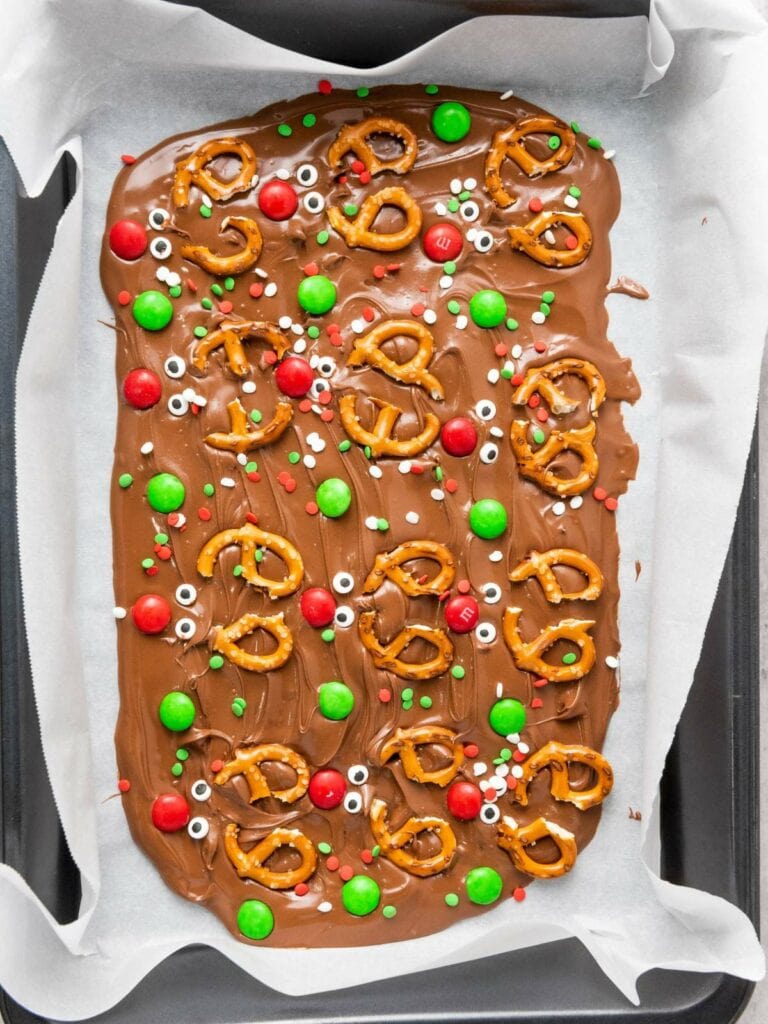 A tray of chocolate bark topped with pretzels, red and green candies, and candy eyeballs on parchment paper.