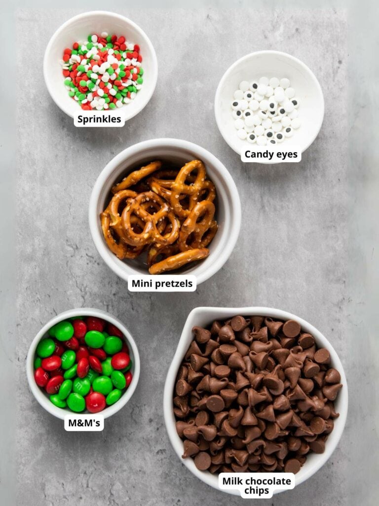 Five bowls containing sprinkles, candy eyes, mini pretzels, M&M's, and milk chocolate chips on a gray surface.