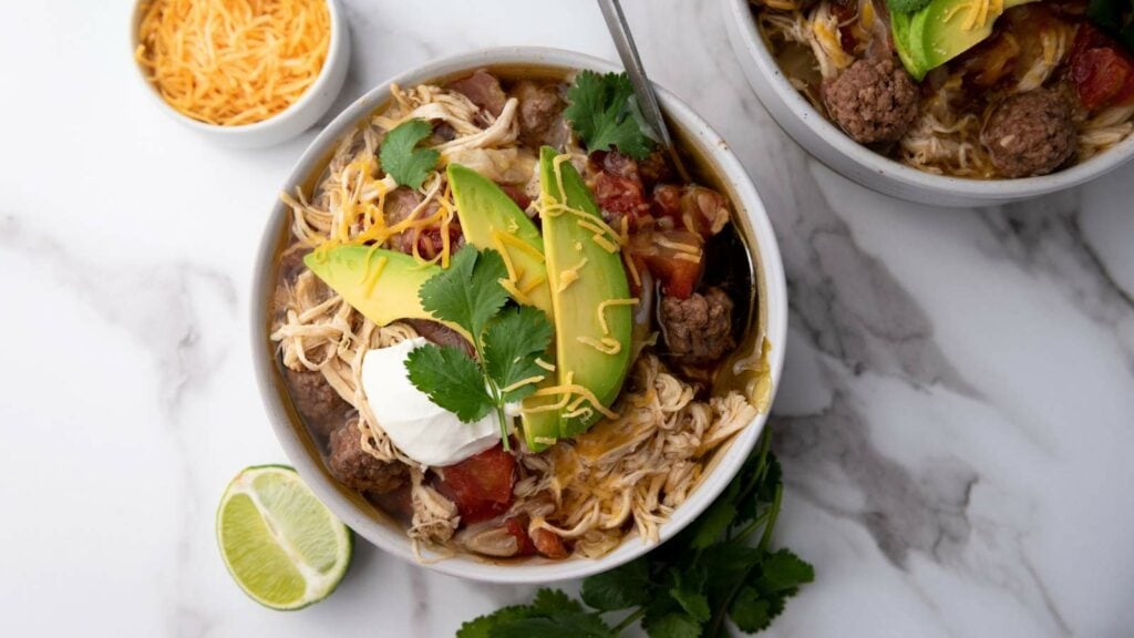 Bowl of taco soup with shredded chicken, avocado, cilantro, cheese, sour cream, and lime on a marble surface.