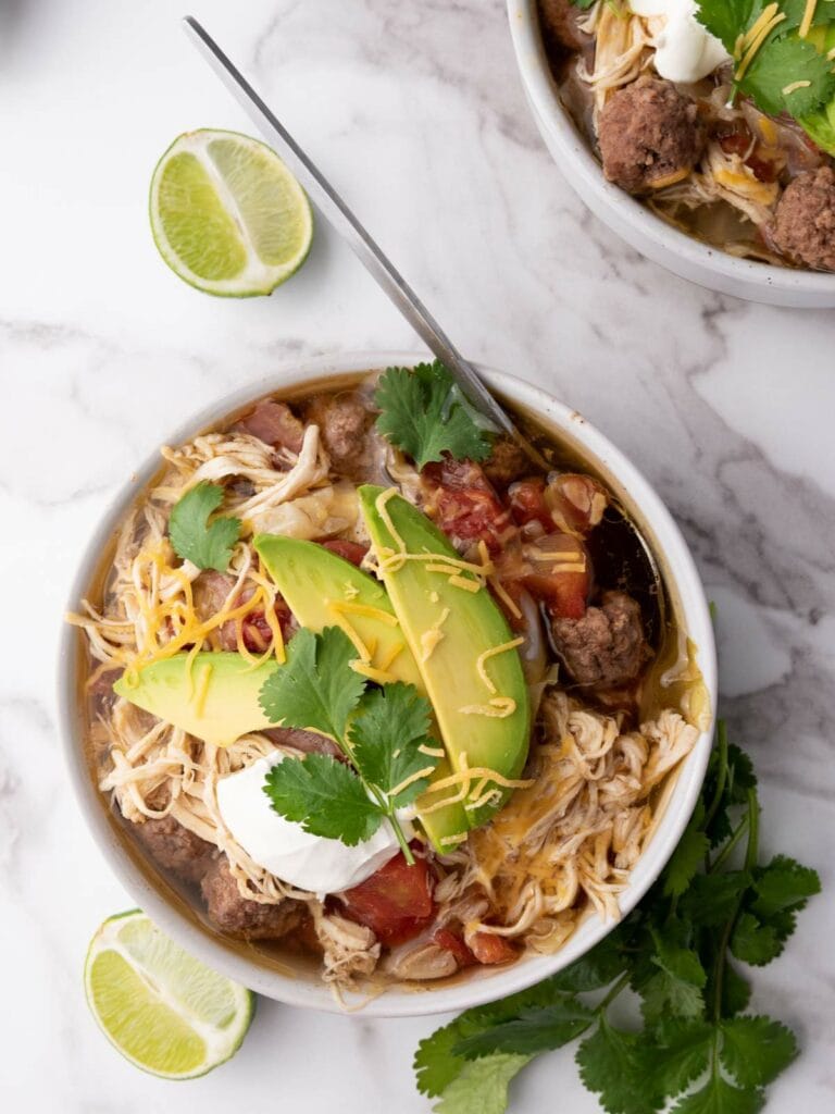 A bowl of taco soup topped with sliced avocado, cilantro, shredded cheese, and sour cream.