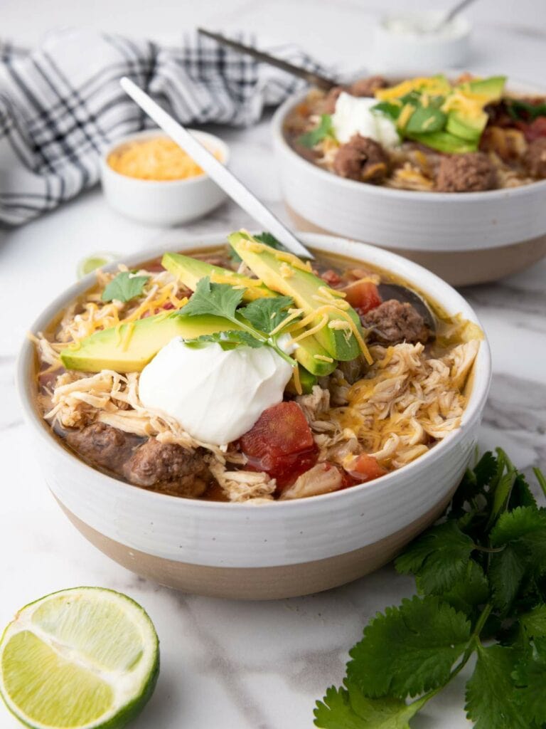 Two bowls of taco soup on a table. A lime half and a small bowl of grated cheese are nearby.