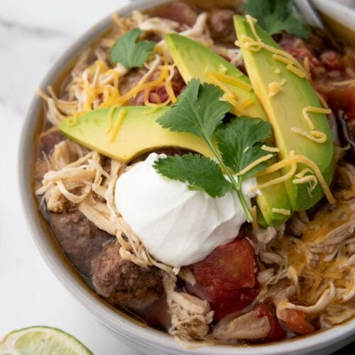 A bowl of taco soup topped with sliced avocado, shredded cheese, cilantro, and a dollop of sour cream.