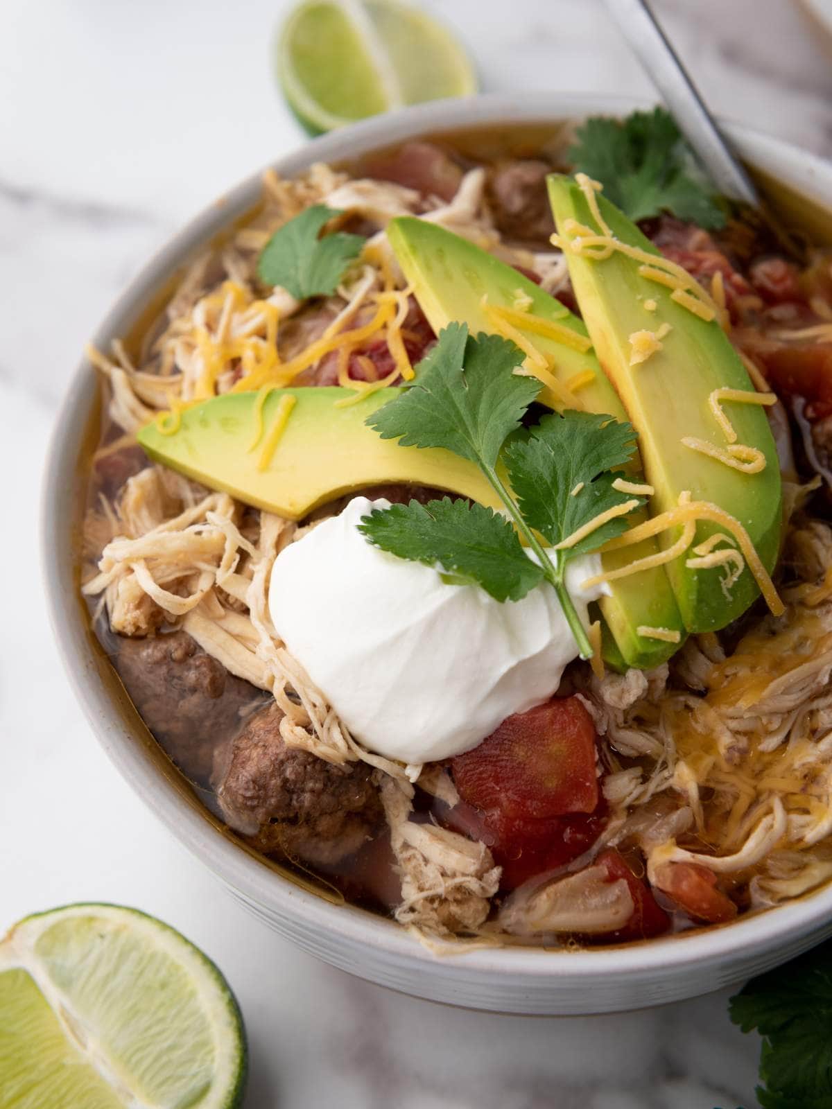 A bowl of taco soup topped with sliced avocado, shredded cheese, cilantro, and a dollop of sour cream.