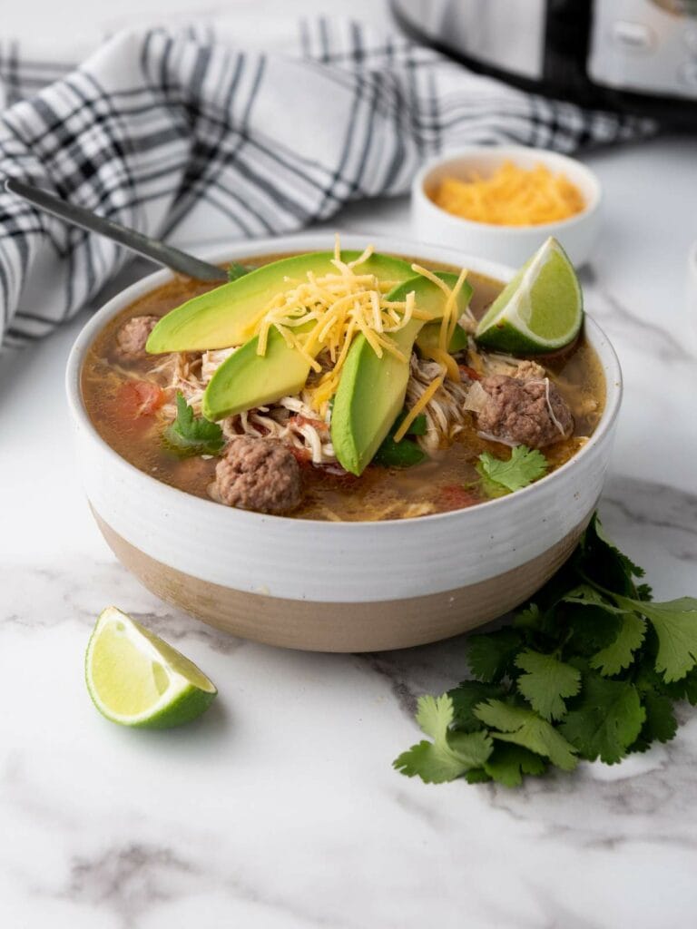 A bowl of taco soup topped with avocado slices, shredded cheese, and meatballs.