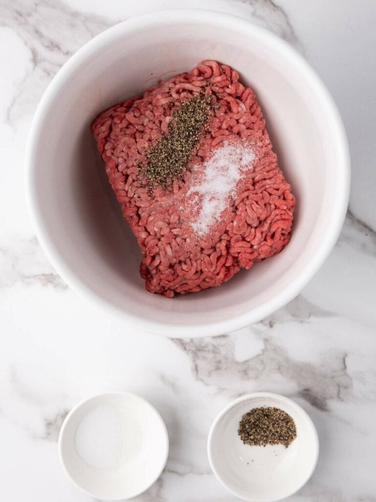 Ground meat in a bowl with salt and pepper, on a marble surface. Two smaller bowls contain additional salt and pepper.