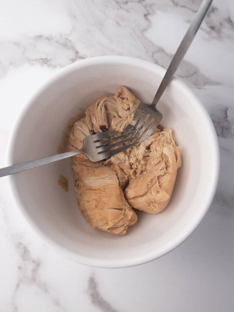 Two forks shred cooked chicken in a white bowl on a marble countertop.