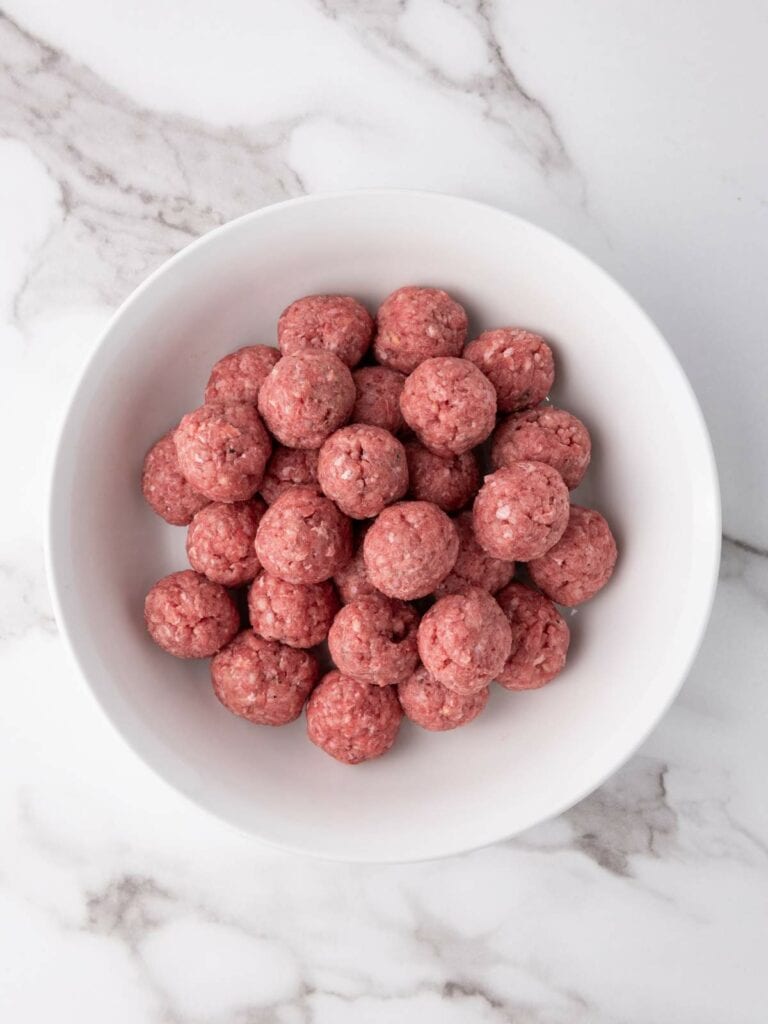 A white bowl filled with raw meatballs on a marble surface.