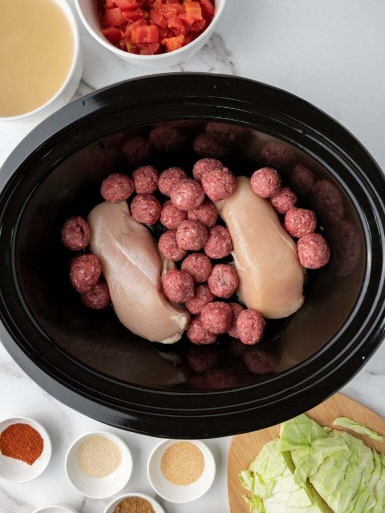 Slow cooker filled with raw chicken breasts and meatballs.