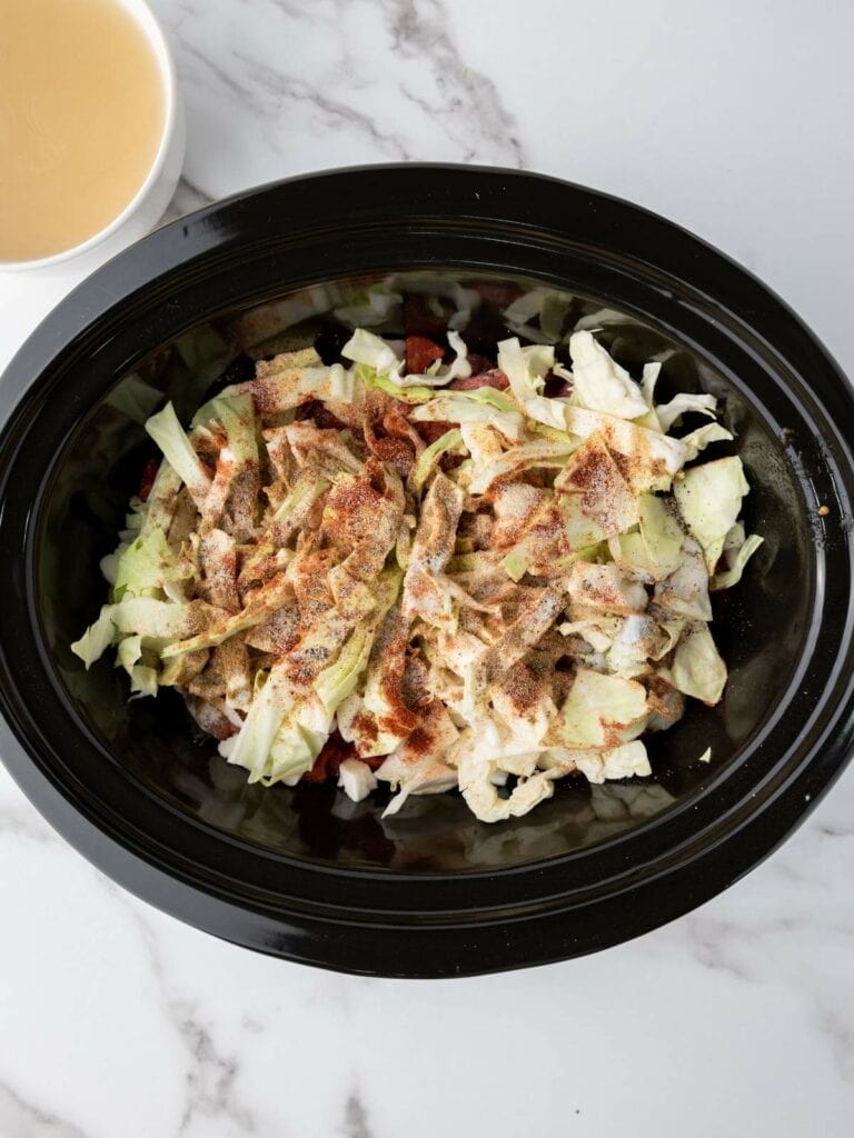 Crockpot filled with cabbage, spices, and other ingredients on a marble surface, next to a bowl of liquid.