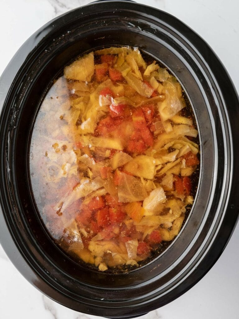 A slow cooker filled with soup containing chopped cabbage, tomatoes, and other vegetables in a clear broth.