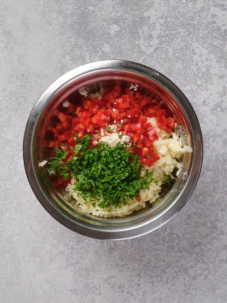 Bowl with chopped red bell peppers, grated cheese, minced garlic, and chopped parsley on a gray surface.