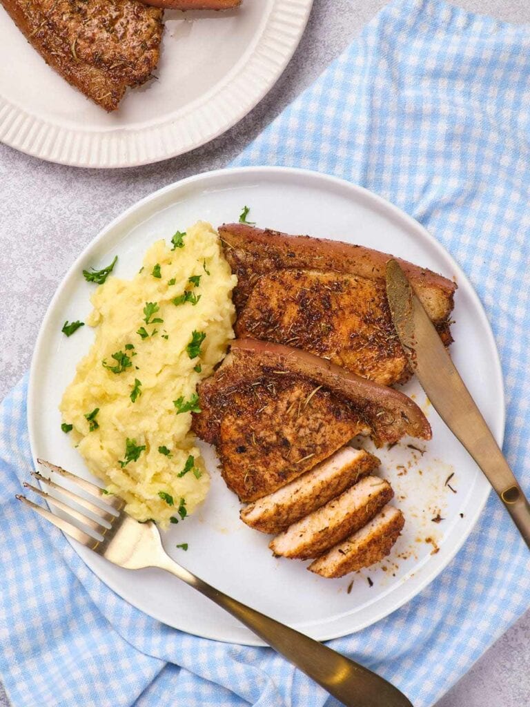 A plate with seasoned pork chops and mashed potatoes garnished with herbs.