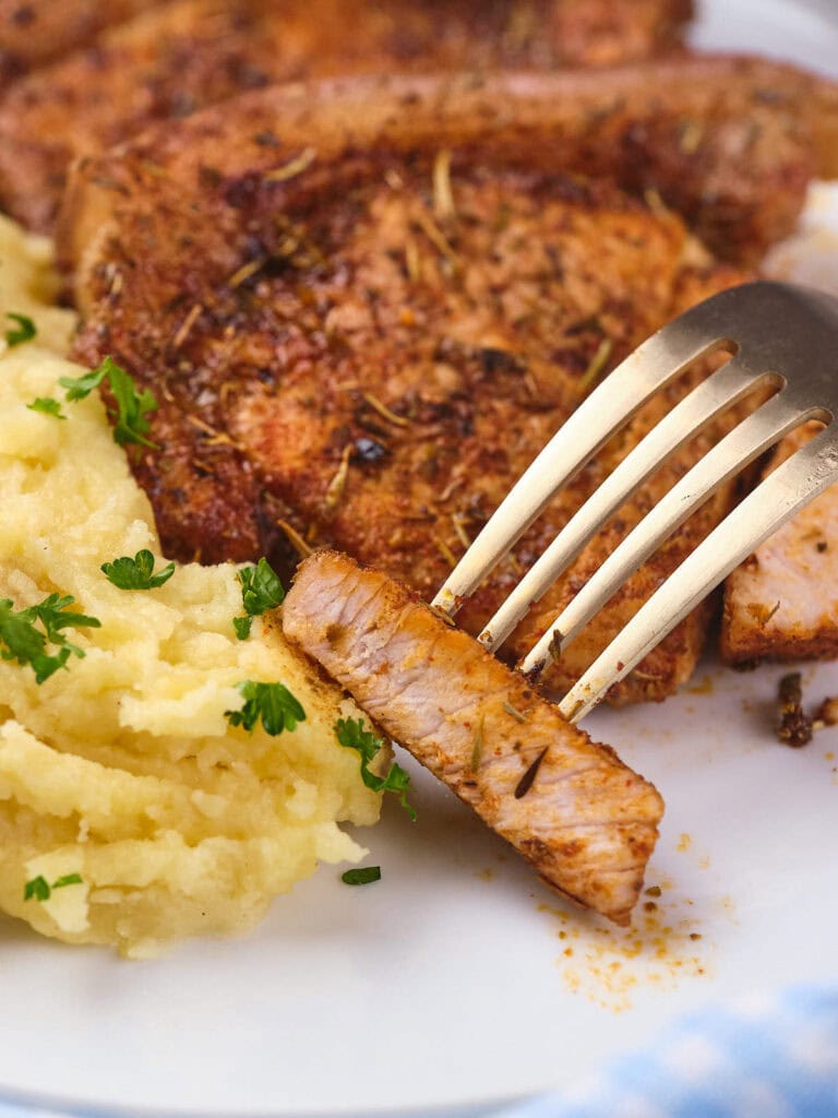 A fork holding a slice of seasoned pork chops beside a serving of creamy mashed potatoes.