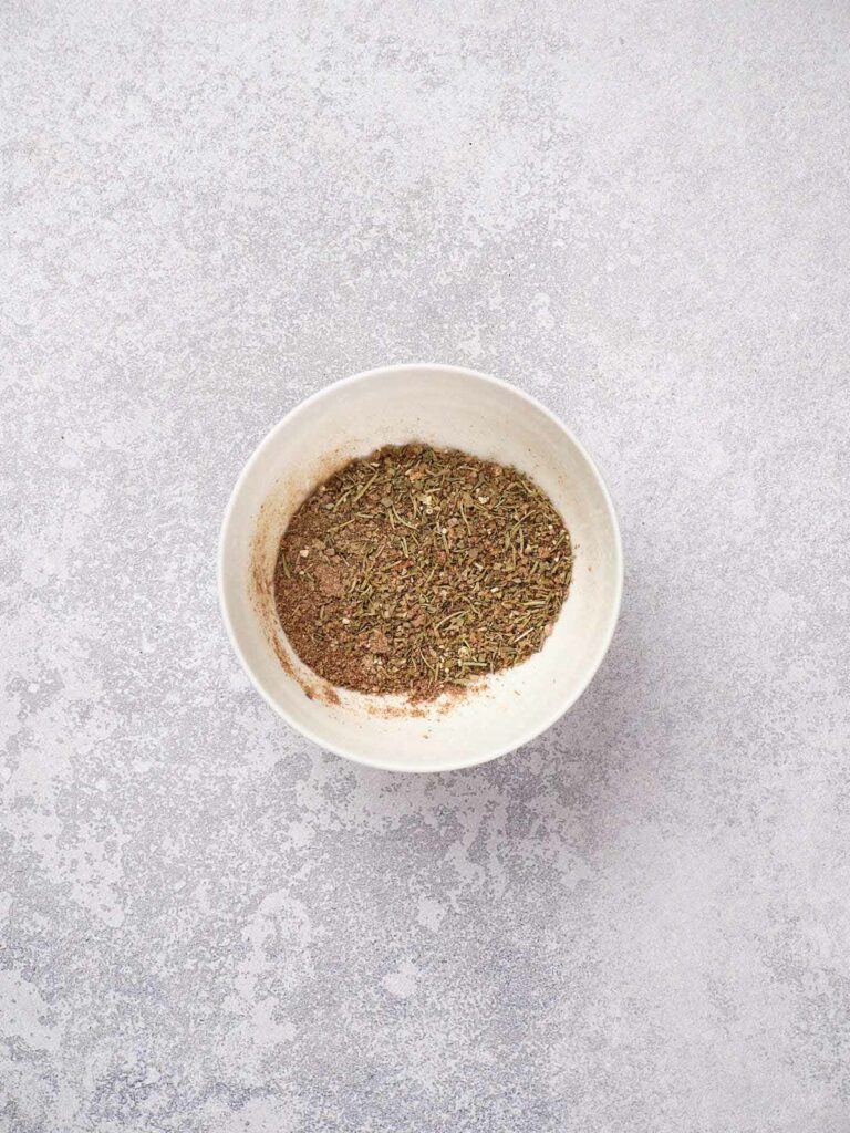 A bowl filled with a mix of dried herbs and spices on a textured gray background.