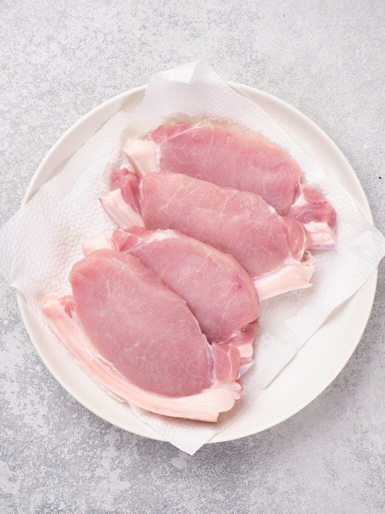 Four raw pork chops on a white plate lined with paper towels, set on a light gray surface.