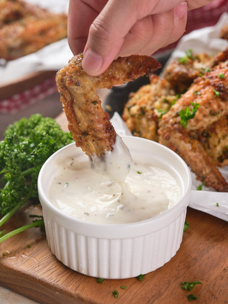 Hand dipping a baked chicken wing into a white sauce in a ramekin.