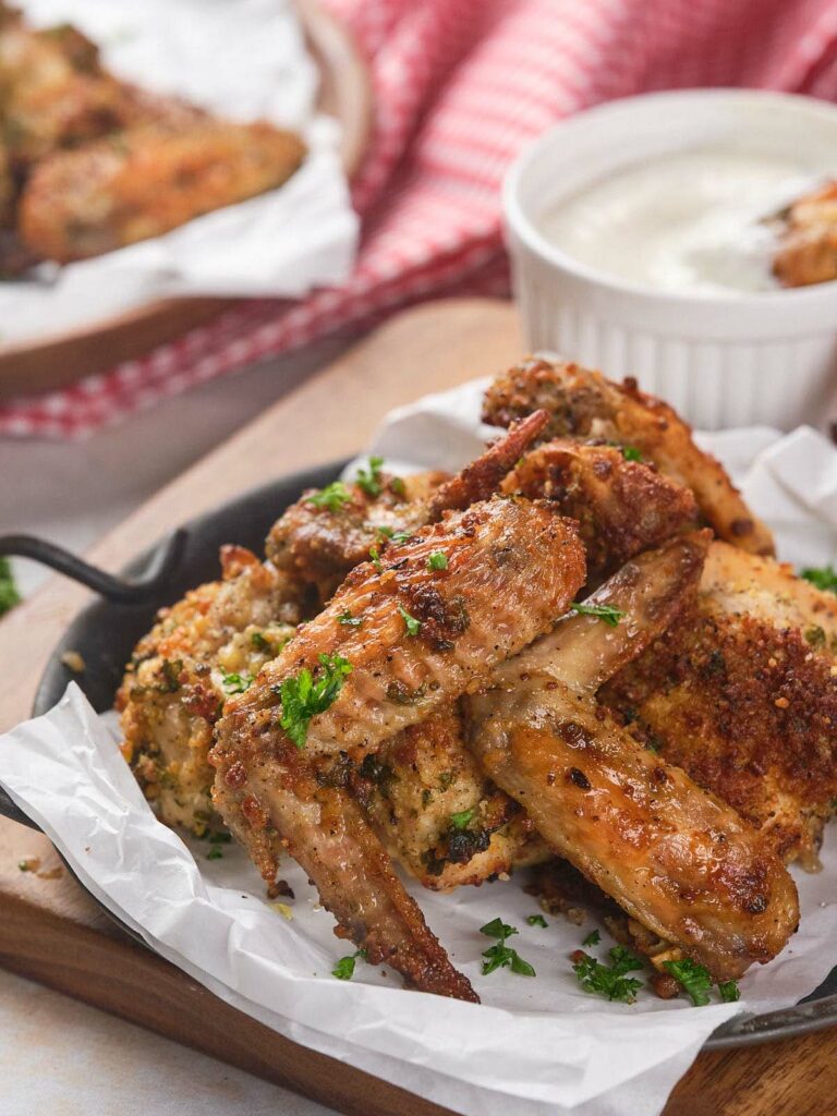 Crispy baked chicken wings garnished with parsley on parchment paper, accompanied by a cup of white dipping sauce.