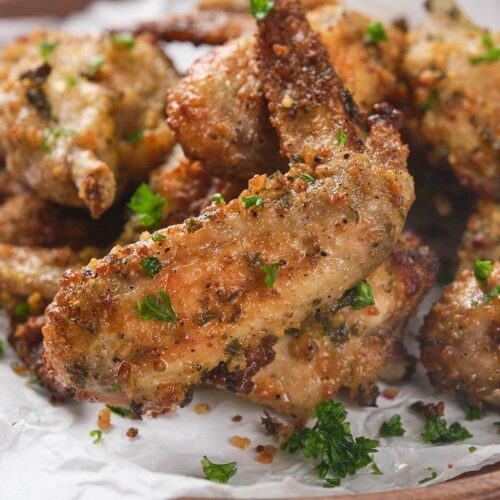 A pile of seasoned, crispy baked chicken wings garnished with chopped parsley on a wooden platter lined with white paper.