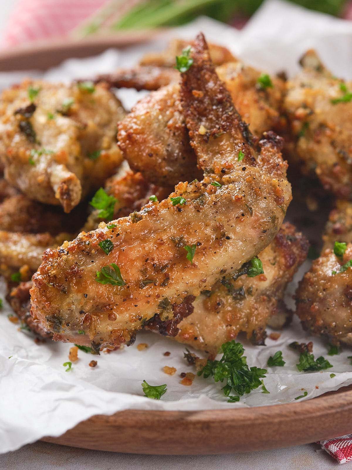 A pile of seasoned, crispy baked chicken wings garnished with chopped parsley on a wooden platter lined with white paper.