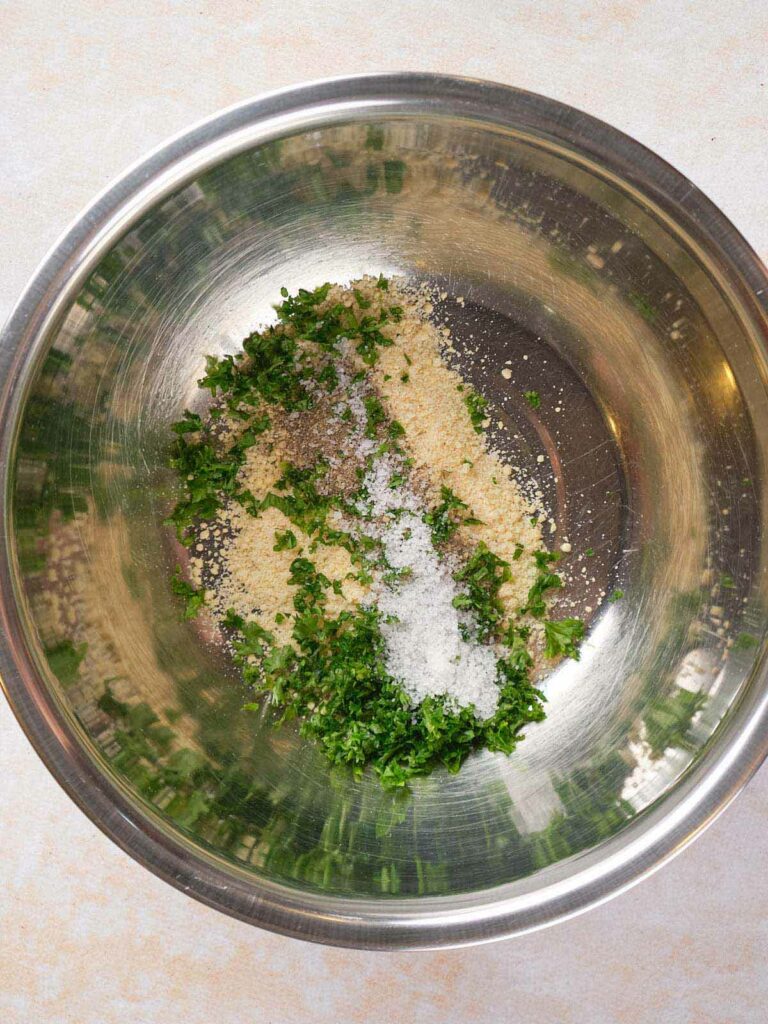 A metal bowl containing chopped herbs, grated cheese, and a mix of salt and pepper.