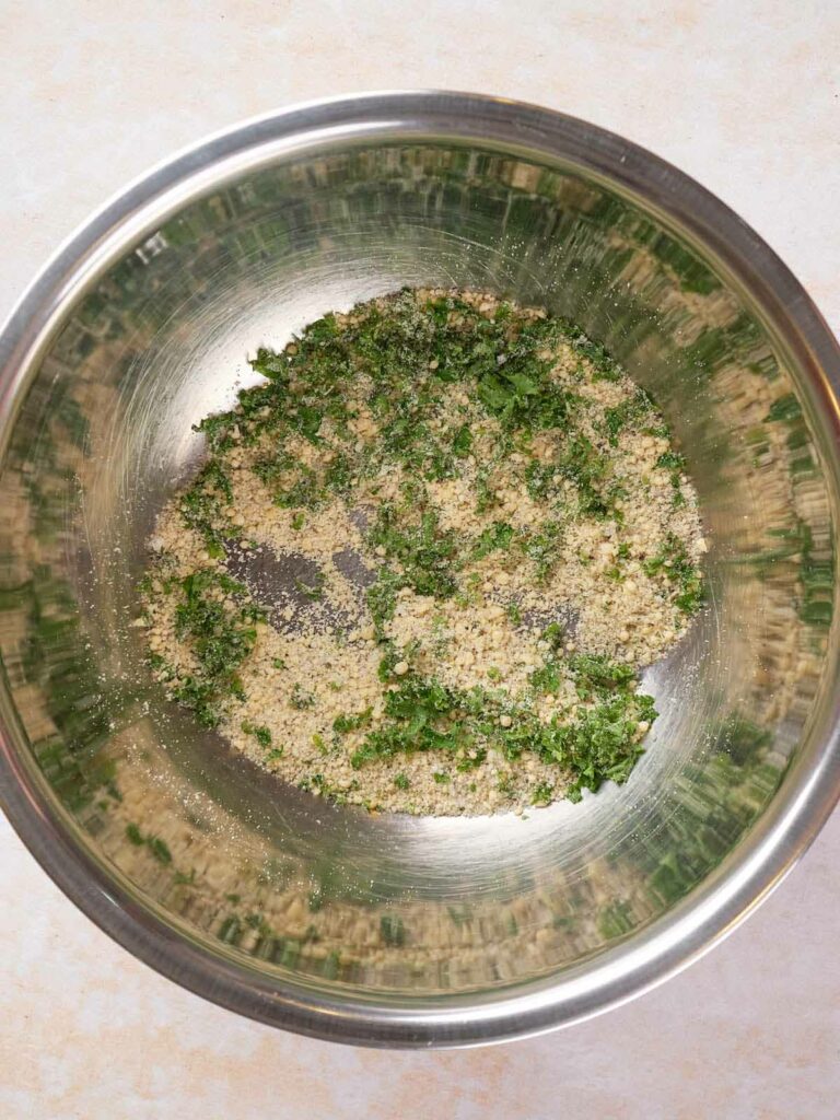 A metal bowl containing a mixture of breadcrumbs and chopped parsley on a light countertop.