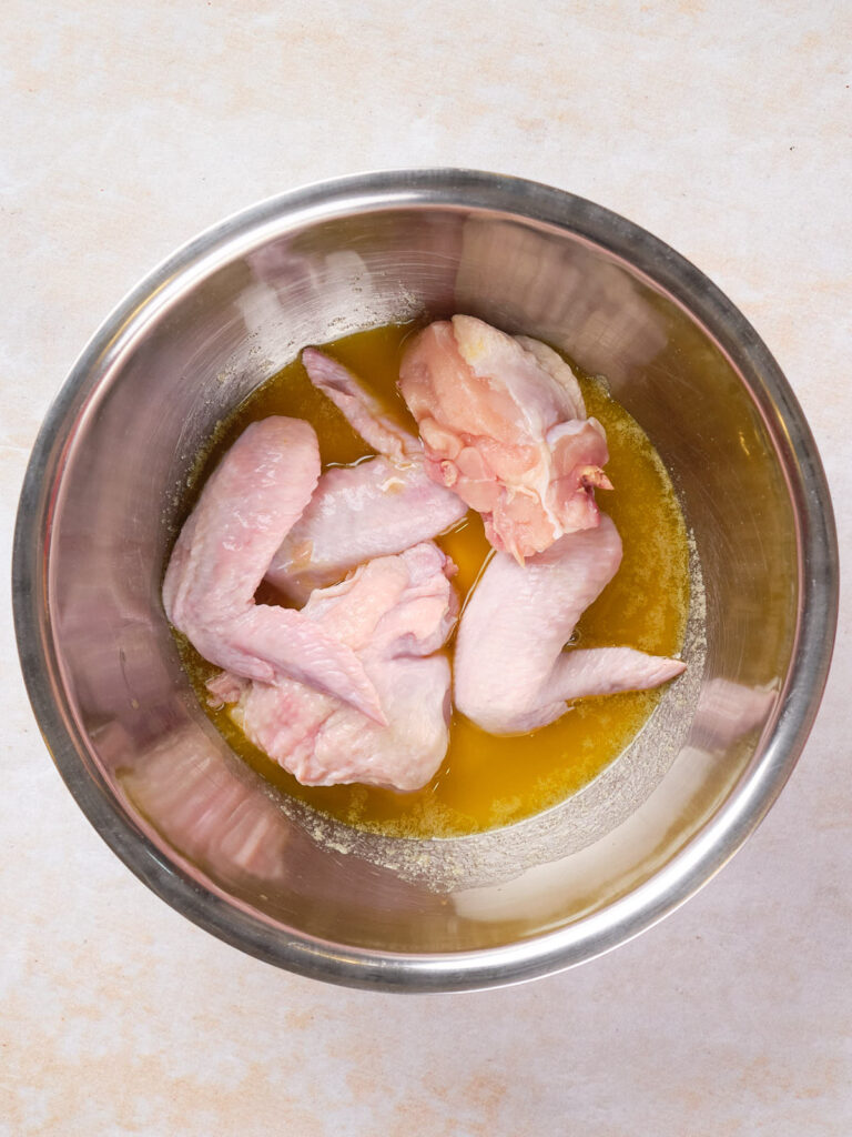 Raw chicken wings in a metal bowl with a yellow marinade on a light-colored surface.