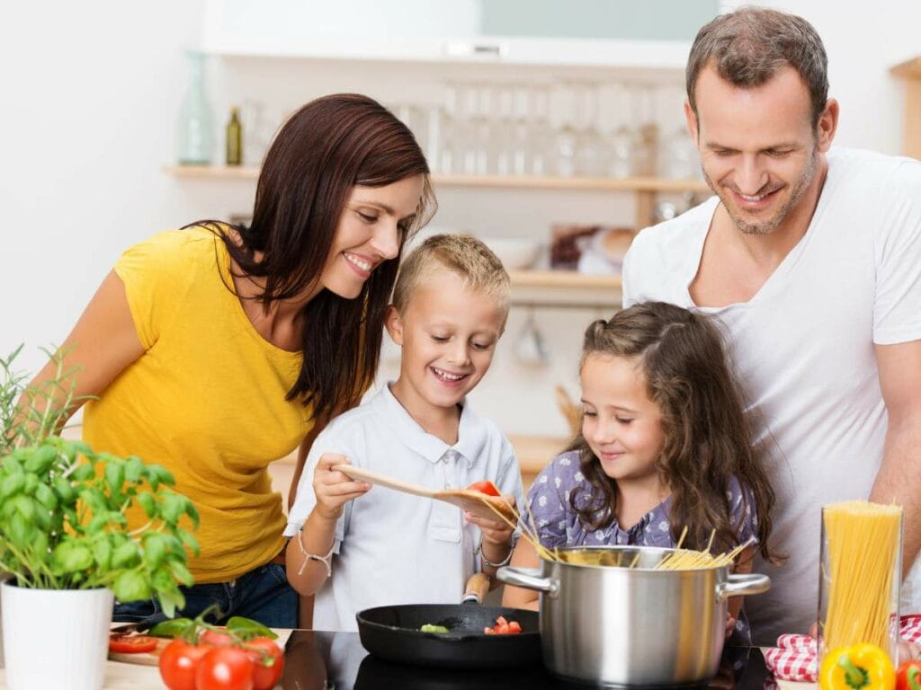 A family of four is cooking together in a kitchen. The children are helping with a wooden spoon and a pot of pasta.