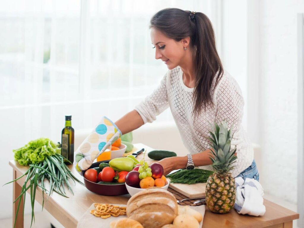 A woman in a kitchen places a pan of vegetables on a wooden table with fresh produce, bread, and olive oil.