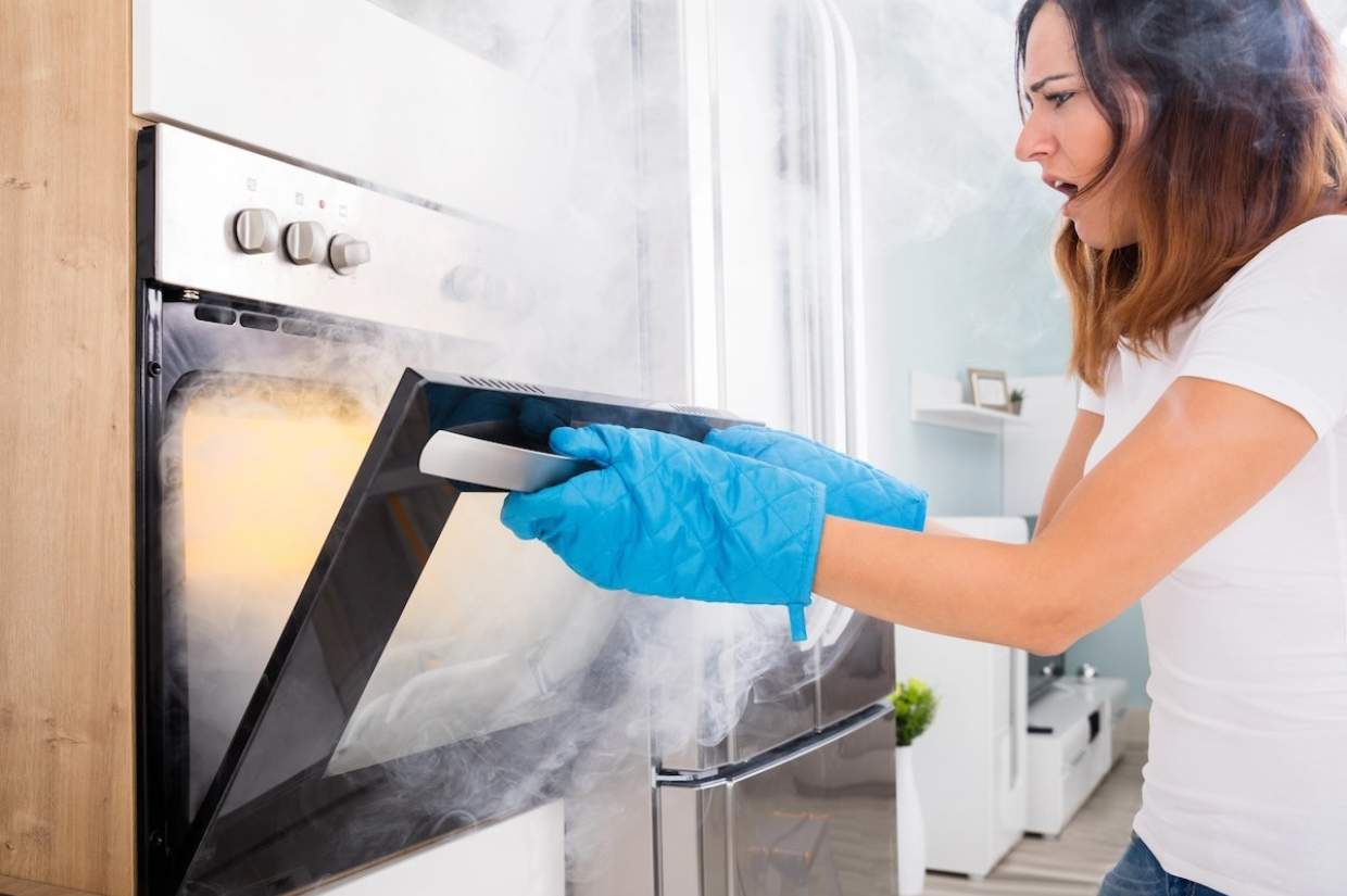 A woman wearing oven mitts opens a smoky oven, looking surprised as she attempts to cook at home.