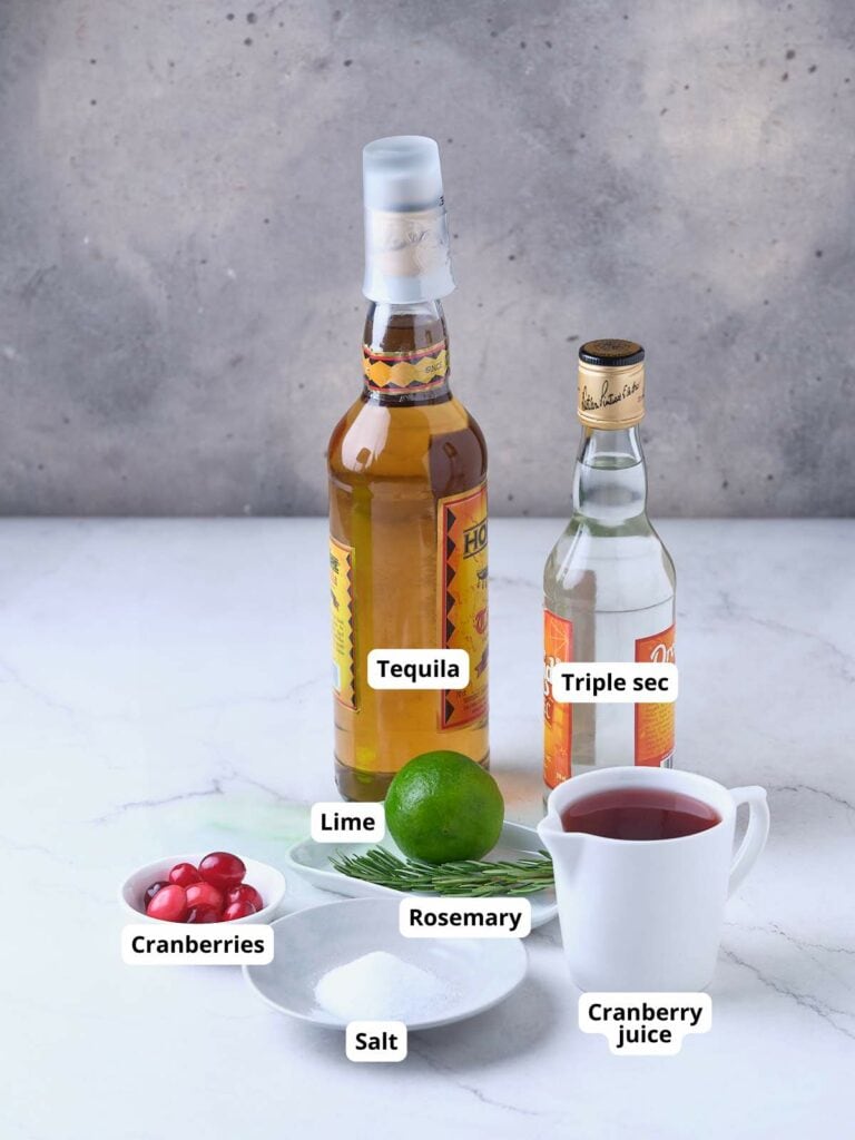 Bottles of tequila and triple sec, with a lime, fresh cranberries, rosemary, salt, and a cup of cranberry juice arranged on a marble surface.