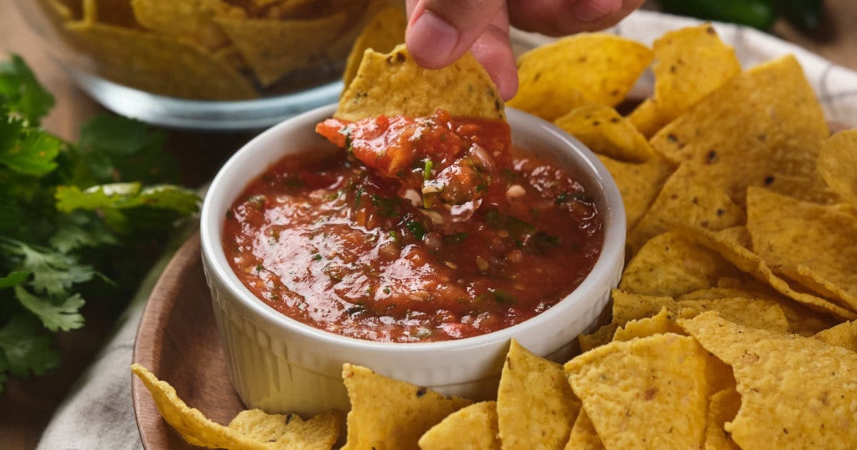 A hand dips a tortilla chip into a bowl of salsa on a plate surrounded by more chips.