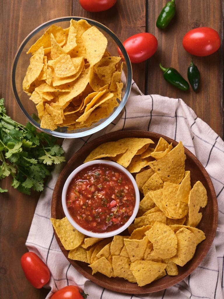 A bowl of tortilla chips and a plate of chips with salsa on a wooden table.