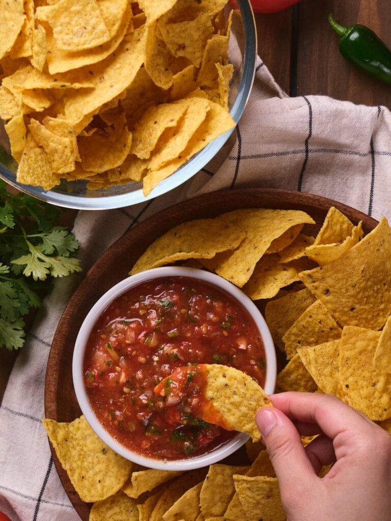 A bowl of salsa surrounded by tortilla chips, with a hand dipping a chip into the salsa.