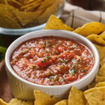 A bowl of tomato salsa surrounded by tortilla chips placed on a wooden plate.