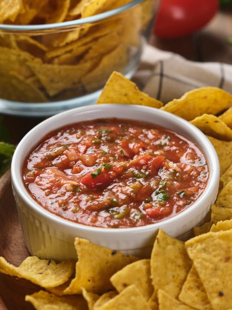 A bowl of tomato salsa surrounded by tortilla chips placed on a wooden plate.